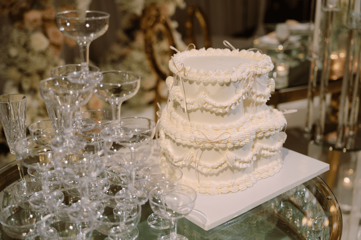 Charming close-up of white wedding cake with pastel pink bows, next to champagne tower, surrounded by glass candle accents