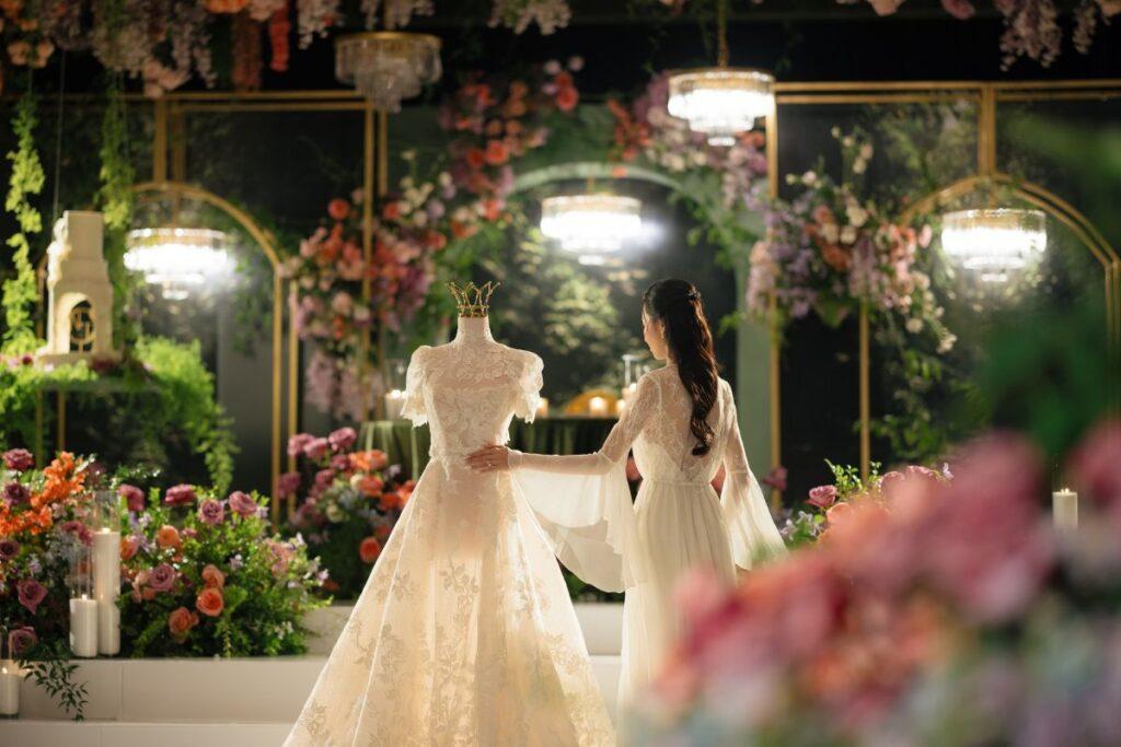 A woman in a flowing white lace gown gently touches a dress form displaying a similar intricate lace wedding dress with a gold crown placed on top. The scene is set in a luxurious, flower-filled venue with vibrant blooms in shades of pink, purple, and orange. Crystal chandeliers hang overhead, reflecting soft light. The lush greenery and floral arrangements in the background create a dreamy, ethereal atmosphere reminiscent of a royal or bridal event.