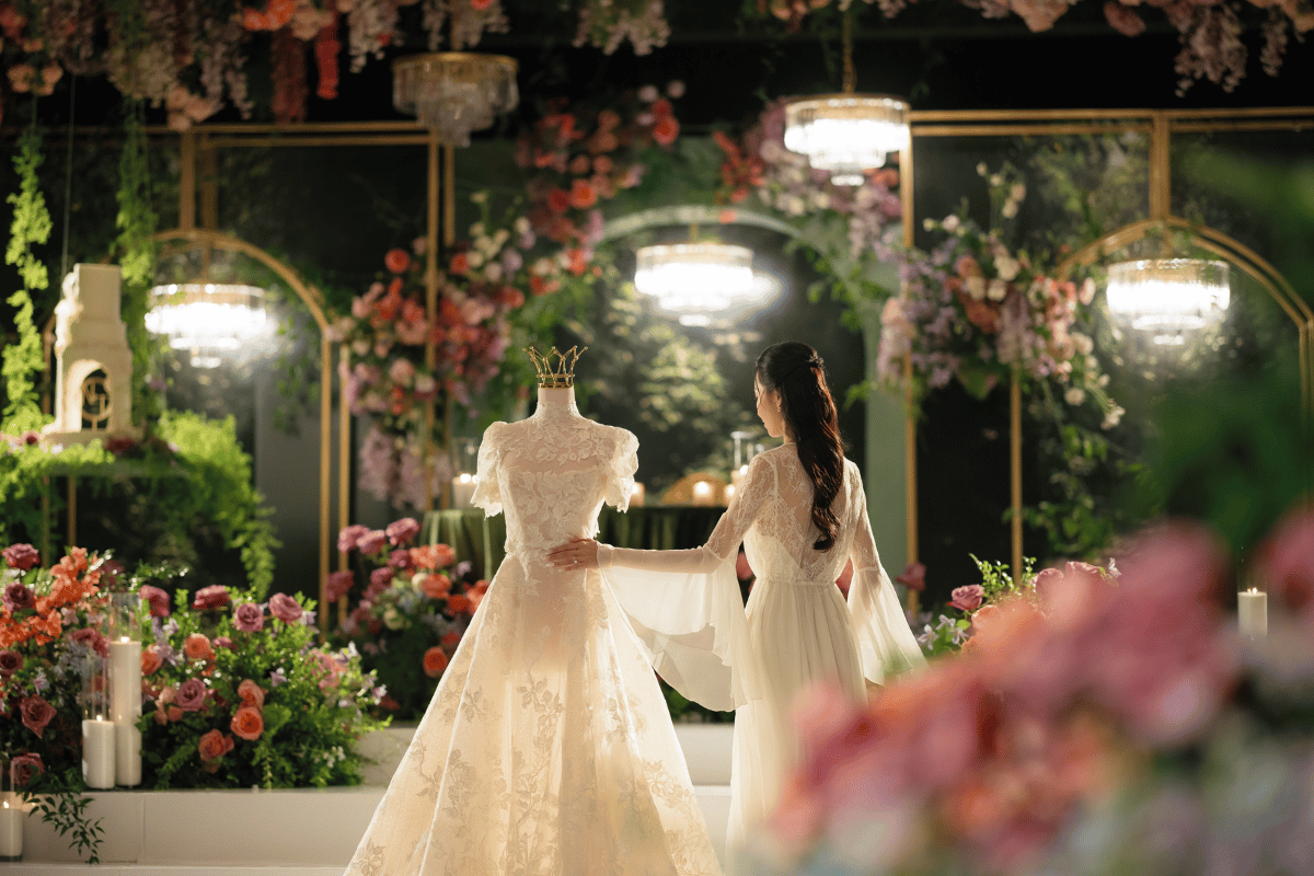 Bride in a lace gown admiring an intricately detailed wedding dress on a mannequin with a crown, surrounded by lush floral arrangements and elegant chandeliers in a romantic setting