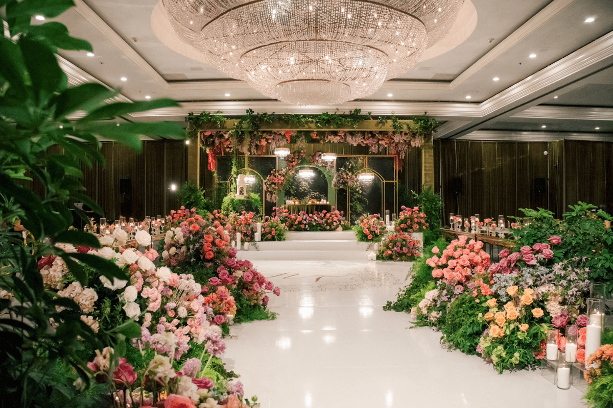 A luxurious wedding venue featuring an elegant crystal chandelier, vibrant floral arrangements in pink, purple, and orange, greenery accents, and candles lining the aisle, creating a romantic and sophisticated atmosphere